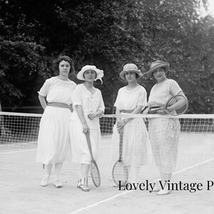 Könnte beinhalten: Vier Frauen in weißen Kleidern und Hüten stehen auf einem Tennisplatz und halten Schläger in der Hand. Sie lächeln alle und schauen in die Kamera. Der Platz ist von einem Netz umgeben und im Hintergrund sind Bäume zu sehen.