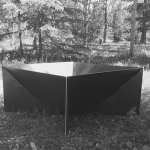 May include: A black metal fire pit with a square shape and four sides. The fire pit is sitting on a bed of gravel and surrounded by green grass.
