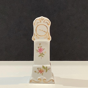 May include: A white ceramic miniature grandfather clock with gold accents and floral details. The clock face is white with gold trim. Pink and orange flowers are painted on the clock's body. The clock is approximately 6 inches tall.