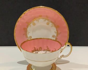 May include: A pink and white teacup and saucer set with gold trim and decorative gold filigree. The teacup is on a small gold stand. The saucer and the inside of the teacup are pink. The set is likely vintage or antique.