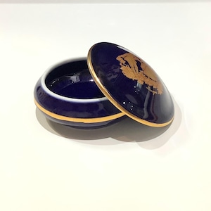 May include: A small, open, navy blue ceramic trinket box with a gold rim and a matching lid. The lid features a gold emblem. The box is on a white surface.