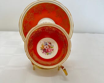 May include: A red teacup and saucer with gold trim and a floral design. The cup and saucer are on a white stand.