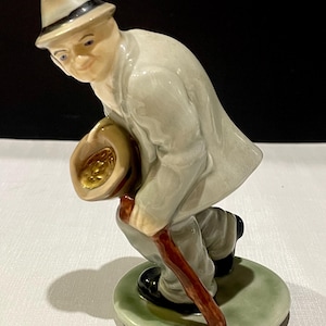 Peut inclure: Figurine en céramique d'un homme en costume gris clair et chapeau crème, tenant un chapeau d'une main et une canne de l'autre. La figurine est sur une base circulaire verte. L'homme est penché, comme s'il marchait.