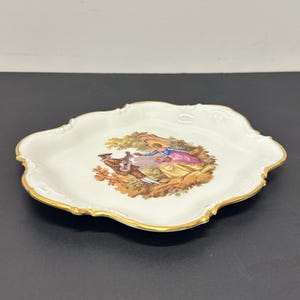 May include: A white porcelain plate with a gold rim features a hand-painted scene of a couple in a garden setting. The man is playing a musical instrument and the woman is holding a bouquet of flowers. The plate has a scalloped edge.