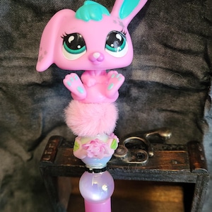 Peut inclure: Stylo rose avec un dessus sur le thème du lapin. Le lapin a un corps rose, des accents bleus et de grands yeux. Sous le lapin se trouve un pompon rose, un motif floral et un globe lumineux. Un accessoire d'écriture amusant et fantaisiste.