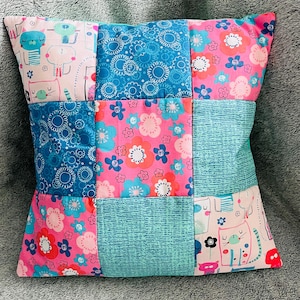 May include: Square patchwork cushion with a variety of patterned fabrics. The cushion features squares of pink floral, blue floral, and teal patterned fabrics. Some squares have cat and animal designs. A decorative home accent.