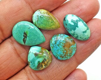 May include: Five polished turquoise gemstones in various shapes, including teardrop and oval. The stones display a range of blue-green hues with brown and black matrix patterns. The gemstones are held in a hand.