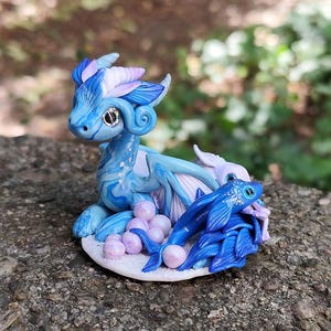 Könnte beinhalten: Handgefertigte Fantasy-Figur eines blauen Drachen mit einem Fisch und lila Kugeln. Der Drache hat einen hellblauen Körper mit dunkelblauen Akzenten und lila und weißen Details an Kopf und Flügeln. Der Fisch ist tiefblau.