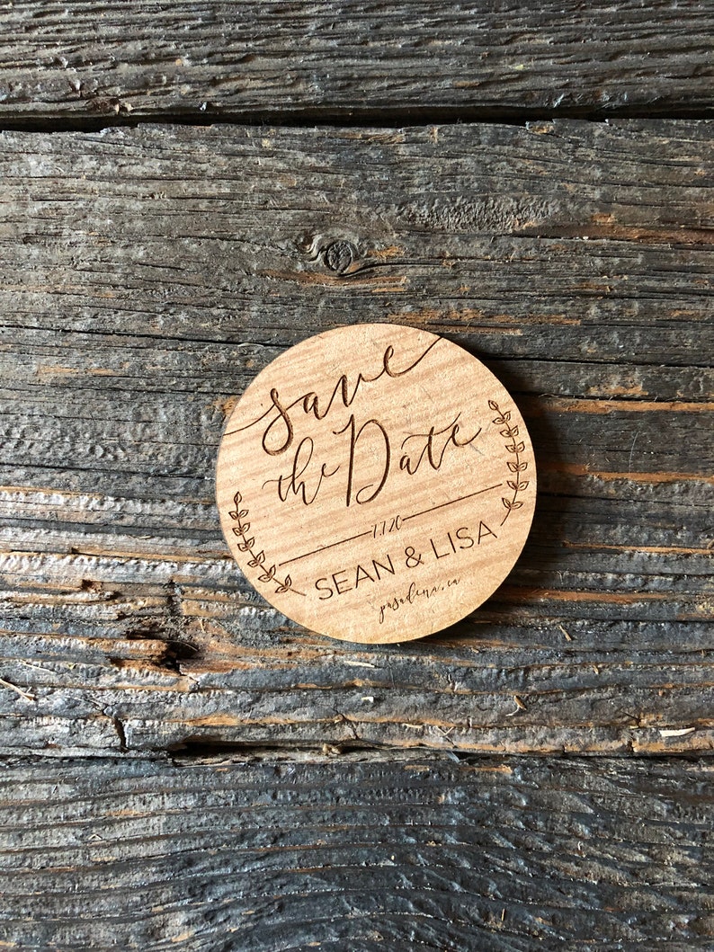 K&ouml;nnte beinhalten: Runde Holz-Ank&uuml;ndigung 'Save the Date'. Der Text 'save the Date' ist in eleganter Schrift gehalten, darunter 'SEAN & LISA'. Die Ank&uuml;ndigung ist von einem Blattdesign umgeben und liegt auf einer verwitterten Holzoberfl&auml;che.