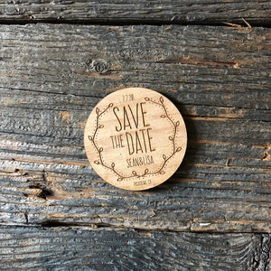 May include: A round wooden save the date sign with a floral border. The text reads "Save the Date" with the date "7.7.20" below. The names "Sean & Lisa" are below the date, and the location "Pasadena, CA" is below the names.