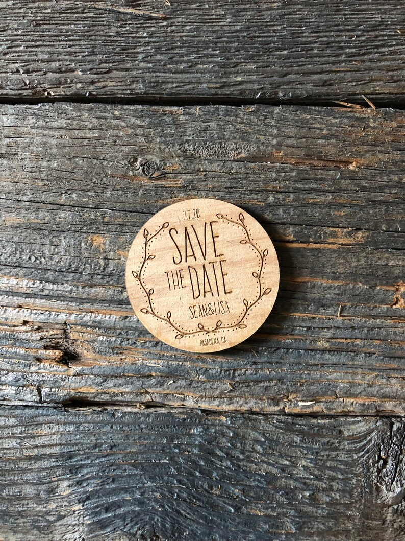 May include: A round wooden save the date sign with a floral border. The text reads "Save the Date" with the date "7.7.20" below. The names "Sean & Lisa" are below the date, and the location "Pasadena, CA" is below the names.