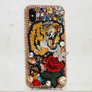Puede incluir: Una funda para iPhone de color oro rosa con un rostro de tigre, una mariposa y una rosa bordados. La funda está cubierta de pedrería y otros adornos.