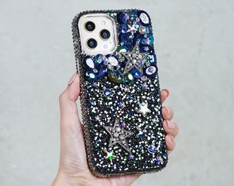 Op de afbeelding: Een zwarte telefoonhoes met een glinsterend, sterrenontwerp. De hoes is versierd met zilveren sterren, blauwe en zilveren strass steentjes en een blauw en zilveren galaxy-achtig patroon.