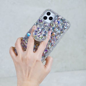 May include: A bedazzled phone case with a matching pop-up grip. The case and grip are covered in iridescent crystals that reflect light. The phone case has a silver frame and a camera cutout.