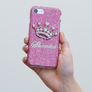 以下が含まれることがあります： ラインストーンで覆われたピンクのiPhoneケース。シルバーのクラウンとラインストーンで「Queen」の文字が飾られています。