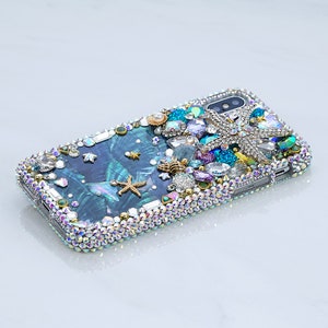 May include: A blue and green iridescent phone case with a mermaid tail, seashells, and starfish design. The case is covered in rhinestones and crystals.
