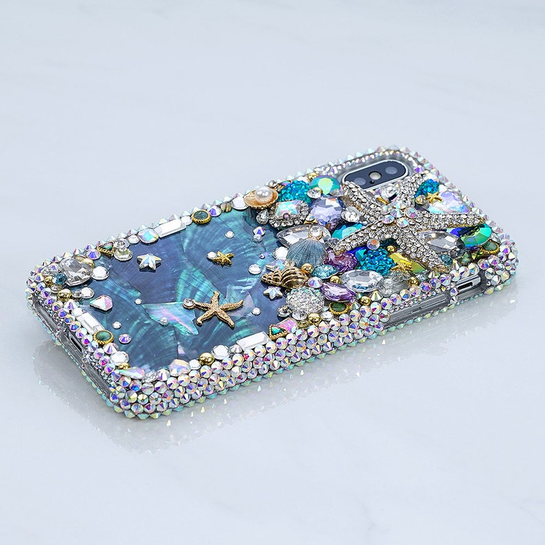 May include: A blue and green iridescent phone case with a mermaid tail, seashells, and starfish design. The case is covered in rhinestones and crystals.