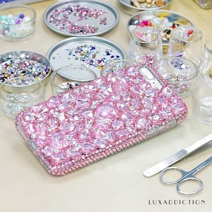 May include: A pink phone case covered in sparkling rhinestones. The case is surrounded by small bowls of loose rhinestones.