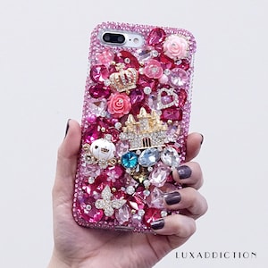 May include: A pink iPhone case covered in rhinestones, crystals, and charms. The case features a pink flower, a crown, a castle, a pumpkin carriage, and a butterfly.
