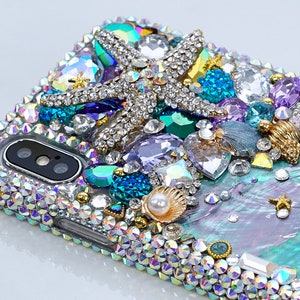 May include: A clear phone case decorated with iridescent rhinestones, a gold starfish, a pearl, and a seashell. The case has a blue, green, and purple iridescent background.