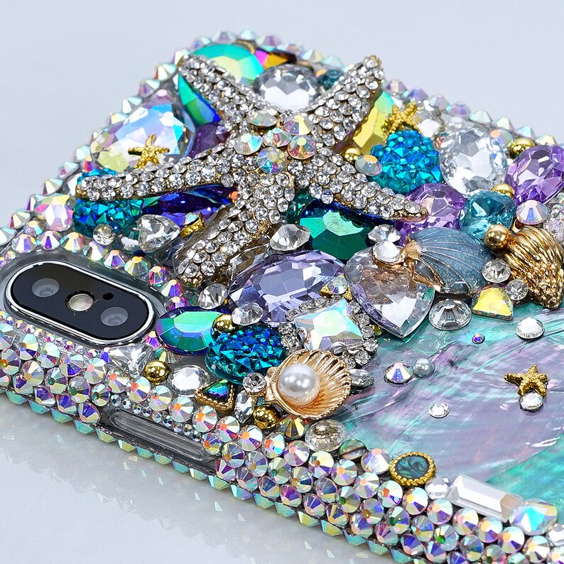 May include: A clear phone case decorated with iridescent rhinestones, a gold starfish, a pearl, and a seashell. The case has a blue, green, and purple iridescent background.