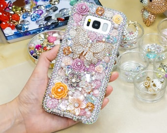 Puede incluir: Una funda de teléfono rosa, blanca y dorada con joyas y un diseño de mariposa. La funda está cubierta de diamantes de imitación y otros adornos decorativos.