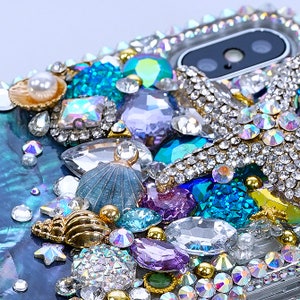 May include: A phone case decorated with a variety of rhinestones, pearls, and seashells. The case features a gold starfish, blue and purple gems, and a white pearl.