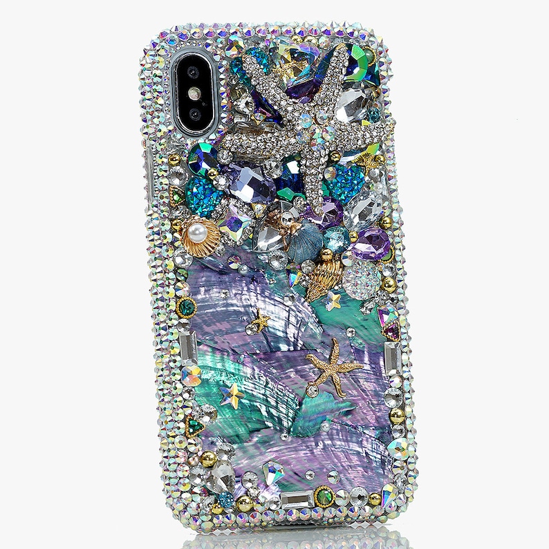 May include: A sparkly iridescent phone case with a mermaid theme. The case is covered in rhinestones, pearls, and seashells. There are two starfish, one large and one small, and a seashell with a pearl inside. The background is a swirling pattern of iridescent blue and purple.