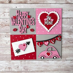 May include: Four wooden squares with Valentine's Day themes. The top left square is gray with the words "Happy Valentine's Day" in pink and red. The top right square is pink with white polka dots and a red heart with the words "Be Mine" in white. The bottom left square is red with white polka dots and a white envelope with a pink heart and arrow. The bottom right square is gray with a pink car with a heart on the hood and a banner that says "LOVE" in white.