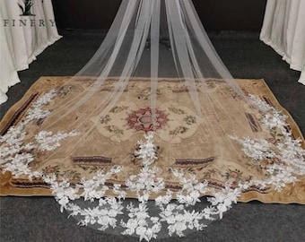 May include: A long, white tulle wedding veil with a delicate floral lace trim. The veil is draped over a rug with a floral pattern.