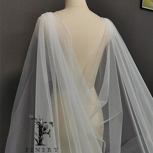May include: A white tulle bridal cape with a long, flowing design. The cape is draped over a dress form and has a V-shaped neckline.