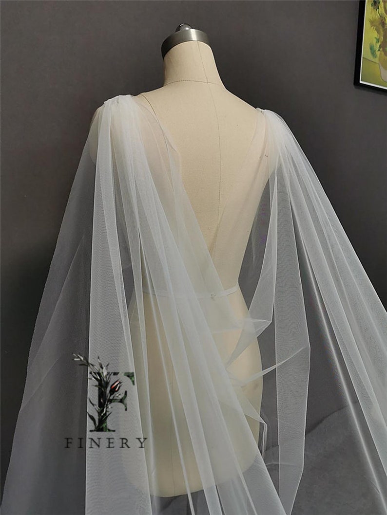 May include: A white tulle bridal cape with a long, flowing design. The cape is draped over a dress form and has a V-shaped neckline.