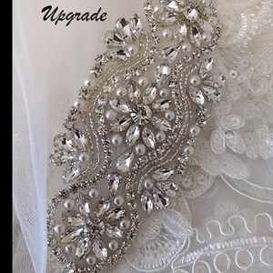 May include: Close-up of a beaded strap embellishment for a wedding dress. The embellishment is made of clear crystals and white pearls, arranged in a floral pattern. The text "Beaded Straps Upgrade" is visible in the top left corner of the image.