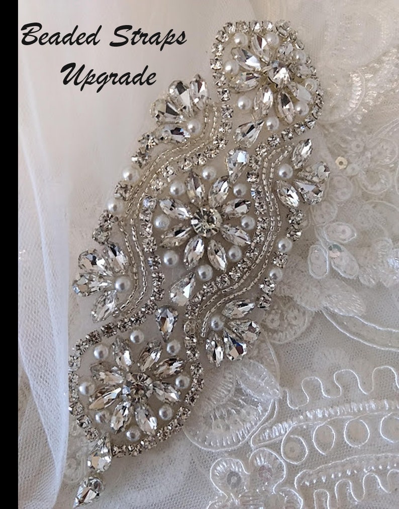 May include: Close-up of a beaded strap embellishment for a wedding dress. The embellishment is made of clear crystals and white pearls, arranged in a floral pattern. The text "Beaded Straps Upgrade" is visible in the top left corner of the image.
