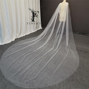 May include: A long, white tulle bridal veil with a cape-like design. The veil is draped over a mannequin and is flowing on the floor.