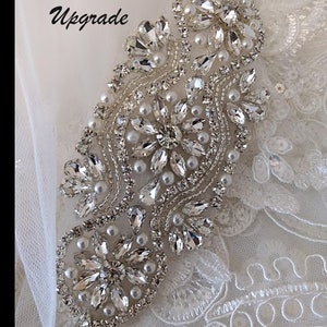 May include: Close-up of a beaded strap embellishment for a wedding dress. The embellishment is made of clear crystals and white pearls, arranged in a floral pattern. The text "Beaded Straps Upgrade" is visible in the top left corner of the image.