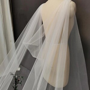 May include: A white tulle bridal cape with a draped design. The cape is made of soft, sheer fabric and has a flowing silhouette.