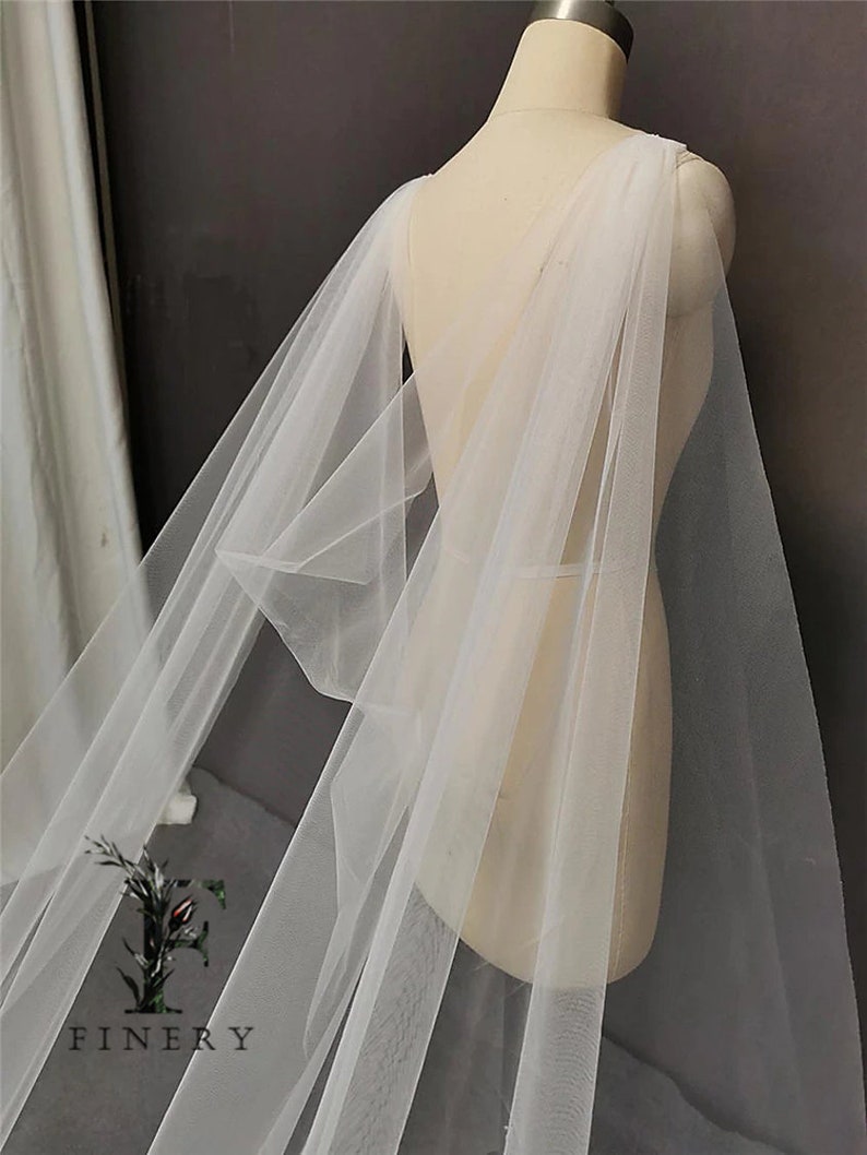 May include: A white tulle bridal cape with a draped design. The cape is made of soft, sheer fabric and has a flowing silhouette.