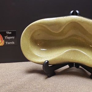 May include: A green ceramic dish with a unique, wavy design. The dish is on a black stand.