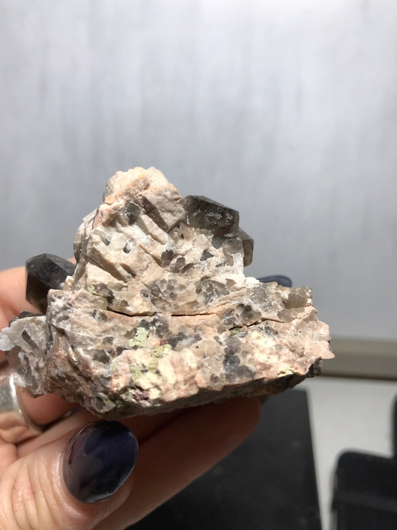 Puede incluir: Un c&uacute;mulo de cristales de cuarzo ahumado incrustados en una matriz rugosa de color gris y marr&oacute;n. Los cristales son transl&uacute;cidos y tienen un color marr&oacute;n ahumado.