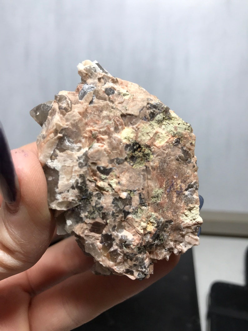Puede incluir: Un cristal &aacute;spero, gris y marr&oacute;n con una punta de cuarzo transparente. El cristal tiene un aspecto natural y bruto y est&aacute; incrustado en una matriz de roca.