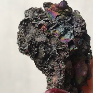Pu&ograve; includere: Primo piano di un minerale grigio scuro iridescente con una superficie ruvida e testurizzata. Il minerale ha un'apparenza metallica e mostra un arcobaleno di colori.