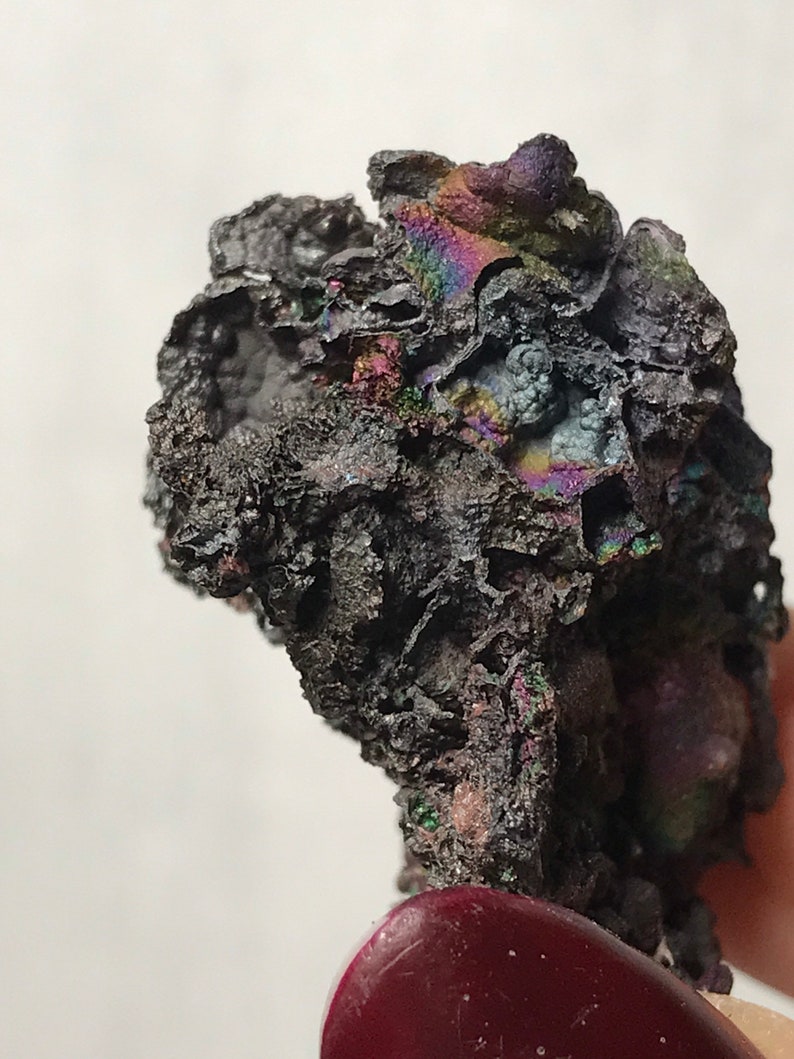Pu&ograve; includere: Primo piano di un minerale grigio scuro iridescente con una superficie ruvida e testurizzata. Il minerale ha un'apparenza metallica e mostra un arcobaleno di colori.