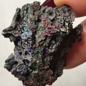 Pu&ograve; includere: Primo piano di un minerale grigio scuro iridescente con una superficie ruvida e testurizzata. Il minerale presenta una variet&agrave; di colori, tra cui viola, verde e blu.