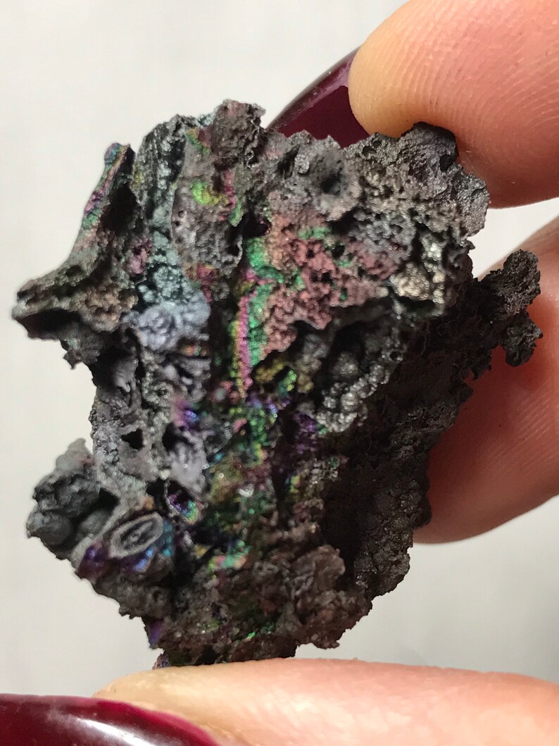 Pu&ograve; includere: Primo piano di un minerale grigio scuro iridescente con una superficie ruvida e testurizzata. Il minerale presenta una variet&agrave; di colori, tra cui viola, verde e blu.