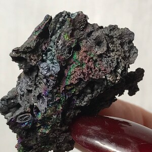 Pu&ograve; includere: Un minerale grigio scuro iridescente con una superficie ruvida e testurizzata. Il minerale ha una lucentezza metallica con sfumature di verde, blu e viola.