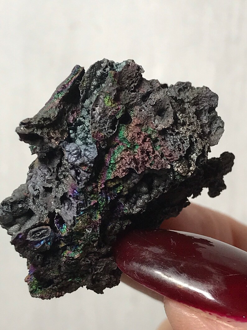 Pu&ograve; includere: Un minerale grigio scuro iridescente con una superficie ruvida e testurizzata. Il minerale ha una lucentezza metallica con sfumature di verde, blu e viola.