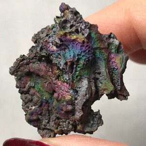 Pu&ograve; includere: Un pezzo di roccia metallica grigia scura iridescente con una superficie ruvida e testurizzata. La roccia ha un bagliore arcobaleno con sfumature di viola, verde e blu.