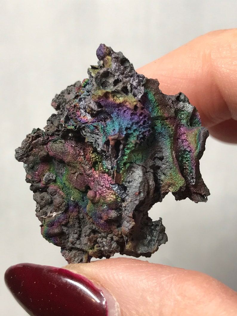 Pu&ograve; includere: Un pezzo di roccia metallica grigia scura iridescente con una superficie ruvida e testurizzata. La roccia ha un bagliore arcobaleno con sfumature di viola, verde e blu.