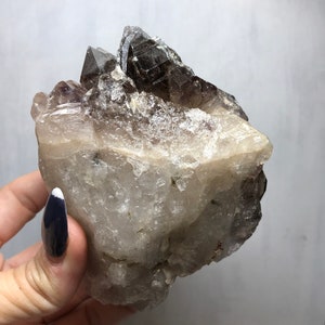 May include: A large, clear quartz crystal cluster with a smoky quartz point on top. The crystal has a natural, rough texture and is a beautiful example of a natural crystal formation.
